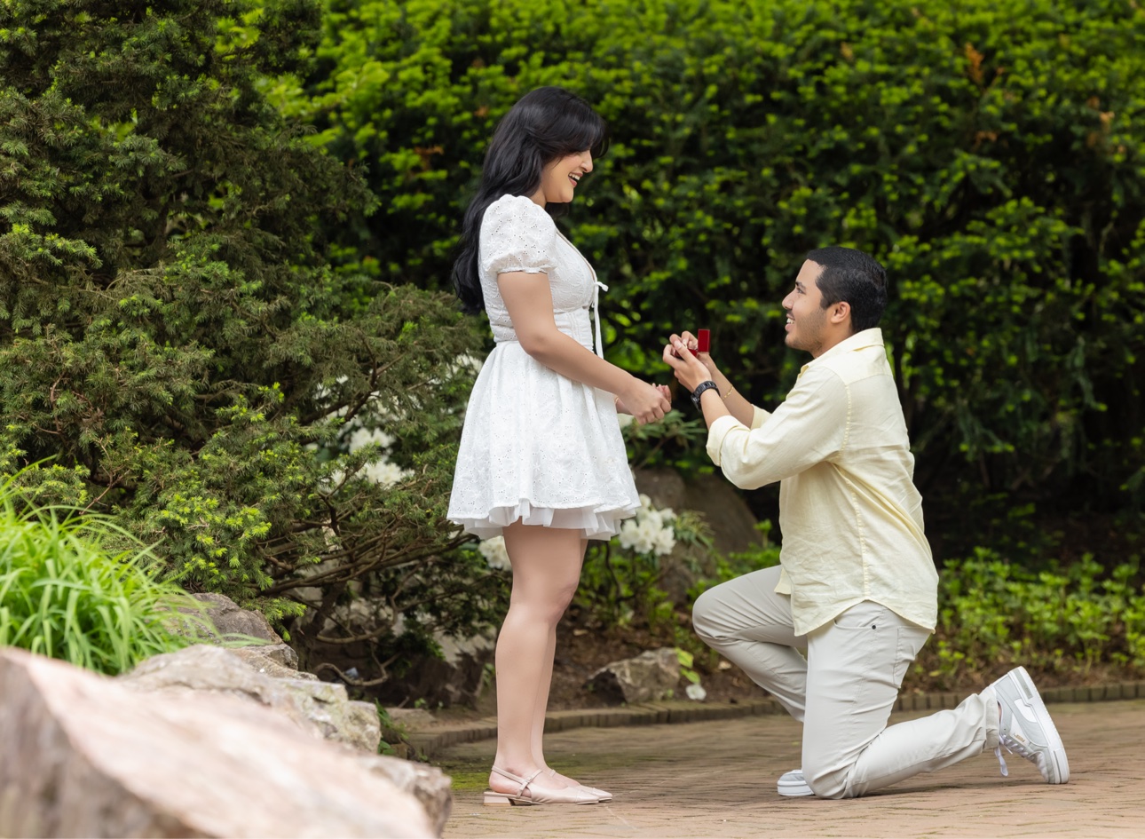 A man kneeling on one knee on a garden path, holding an open red ring box toward the woman he loves as she smiles down at him