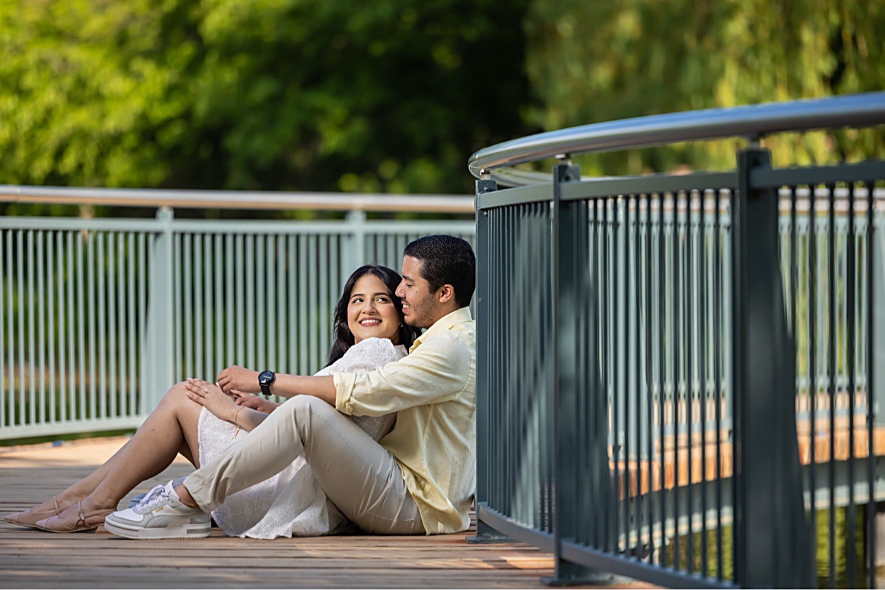 The couple sitting together on the wooden deck of a curved garden bridge, his arm around her as they smile in warm afternoon light
