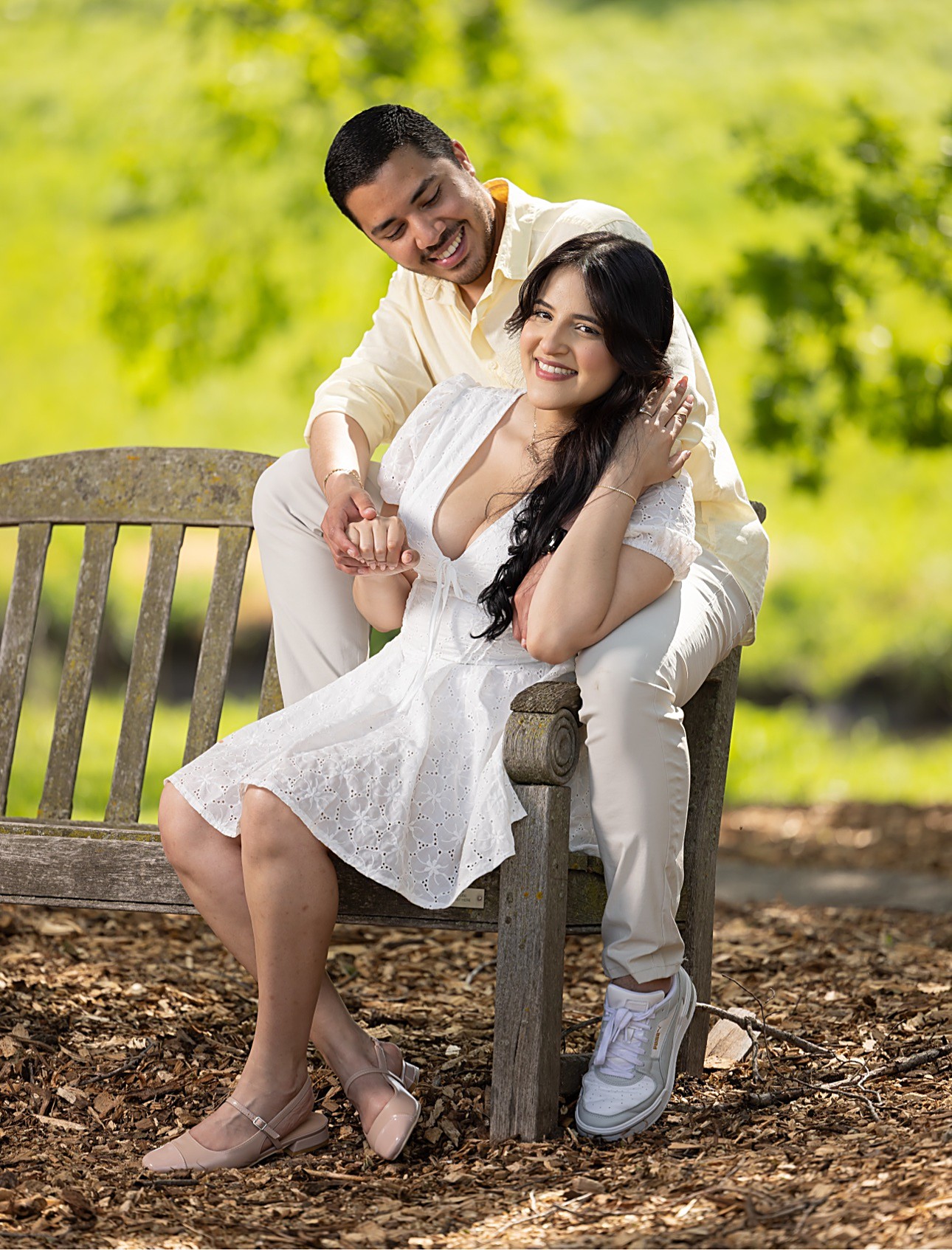A closer portrait of the couple laughing on a garden bench, her hand with the new engagement ring resting over his