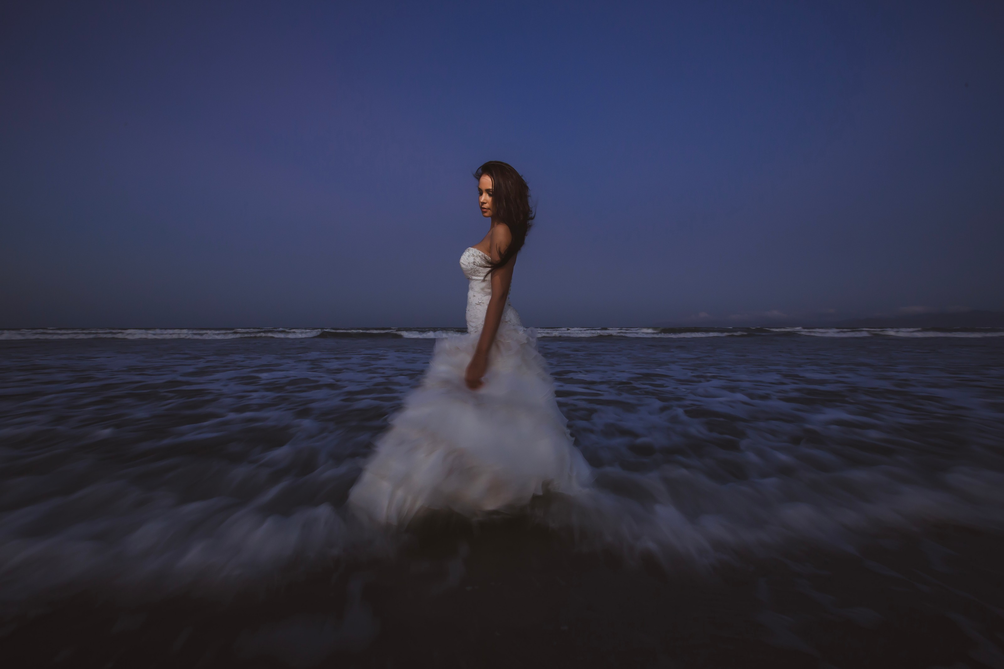 Bride in a white gown standing in the ocean surf at dusk, looking over her shoulder beneath a deep blue sky
