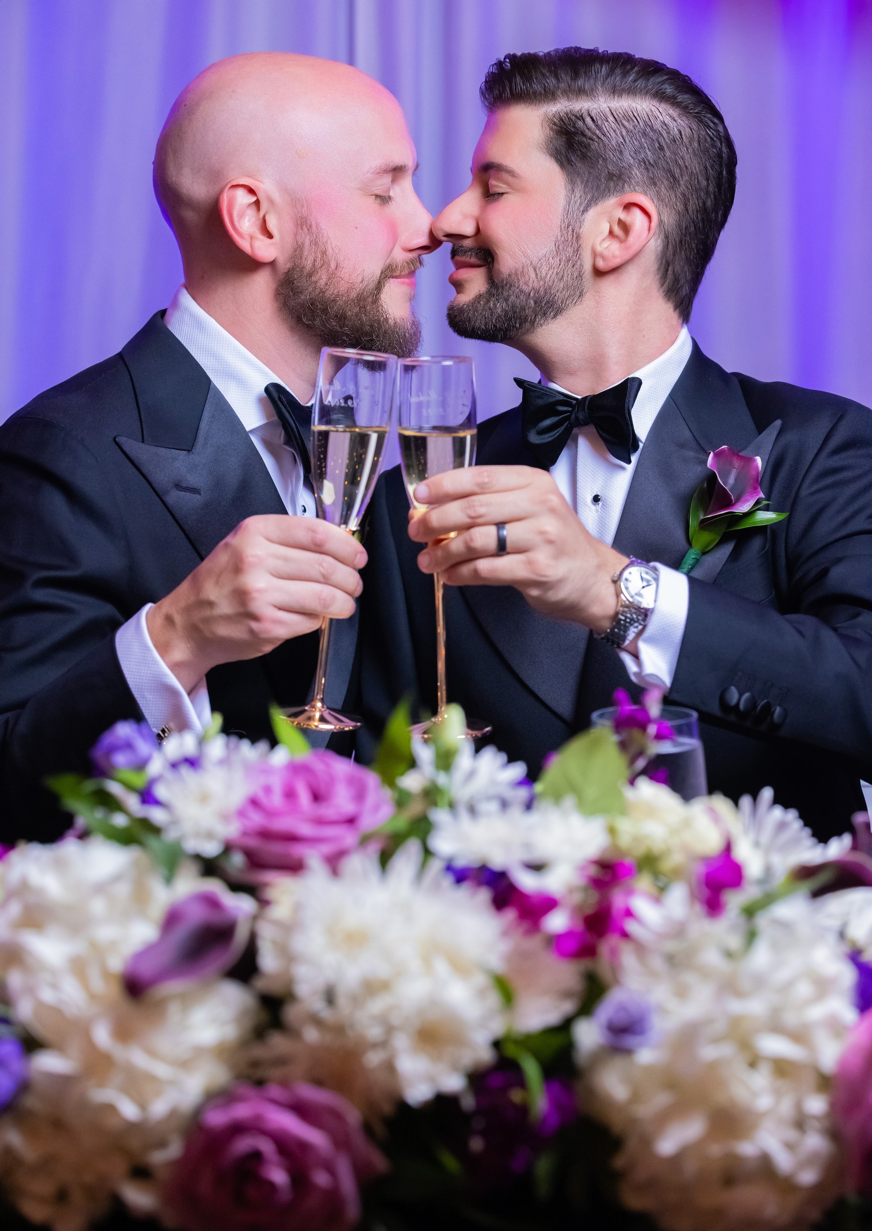 Michael and Kevin touching foreheads and laughing at their sweetheart table, champagne glasses and flowers before them